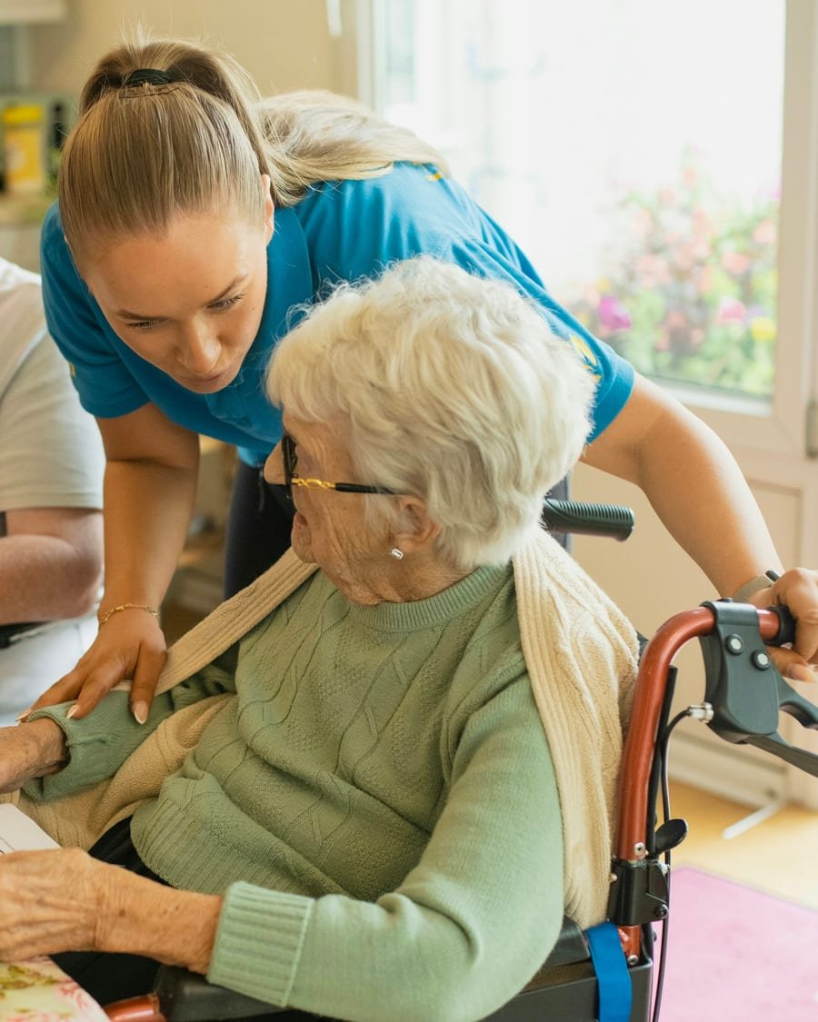 A home health nurse in uniform attends to a seated older adult at a bright kitchen table.