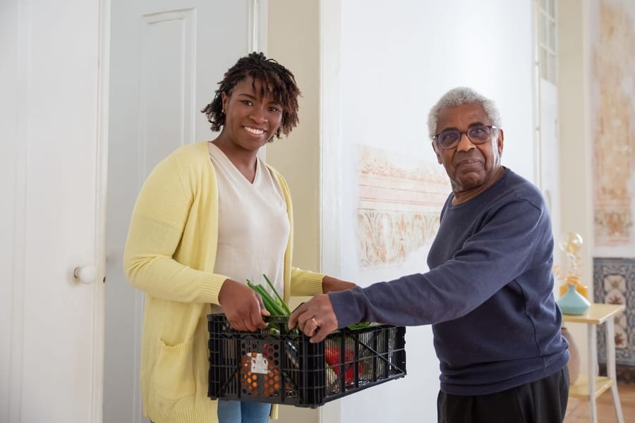 A caregiver brings a box of fresh groceries to an older adult at the door of his home.