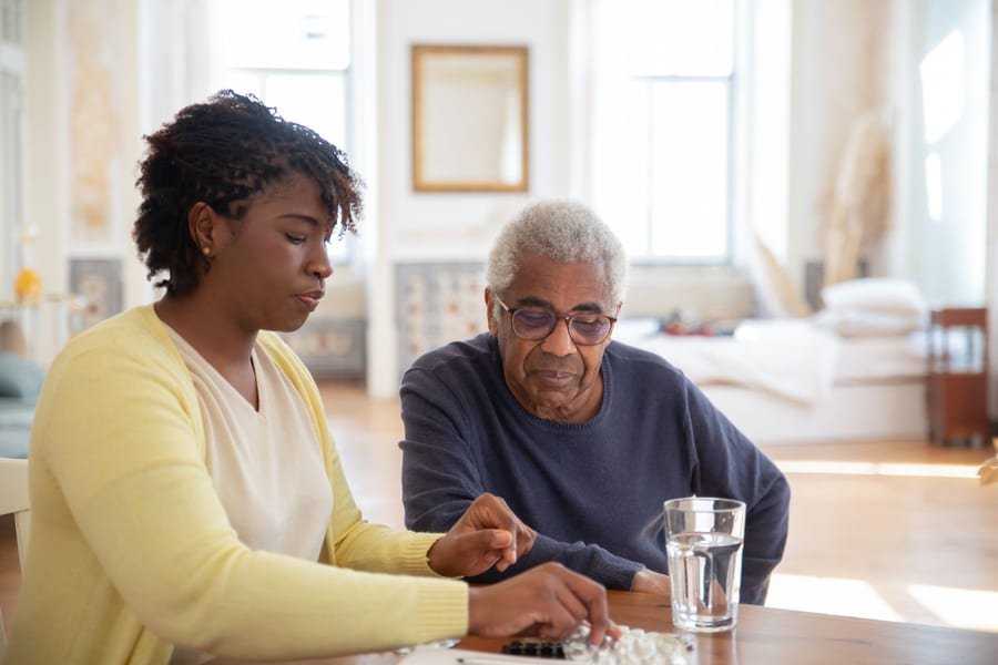 A home health aide helps an older adult organize daily medications at a bright kitchen table.